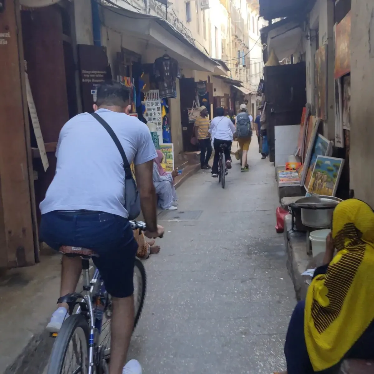 Cyclists riding through a narrow lane of shops and paintings