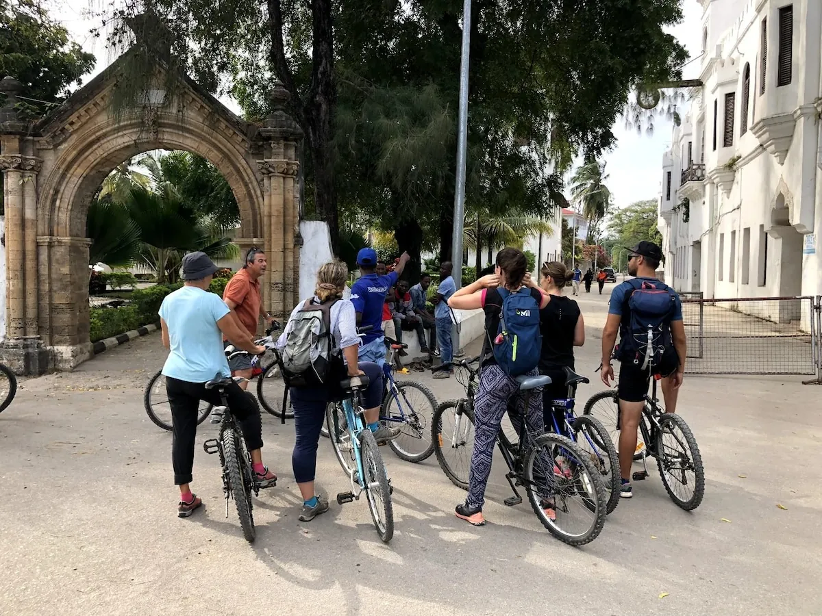 Group of cyclists with bikes beside a carved stone arch and white buildings