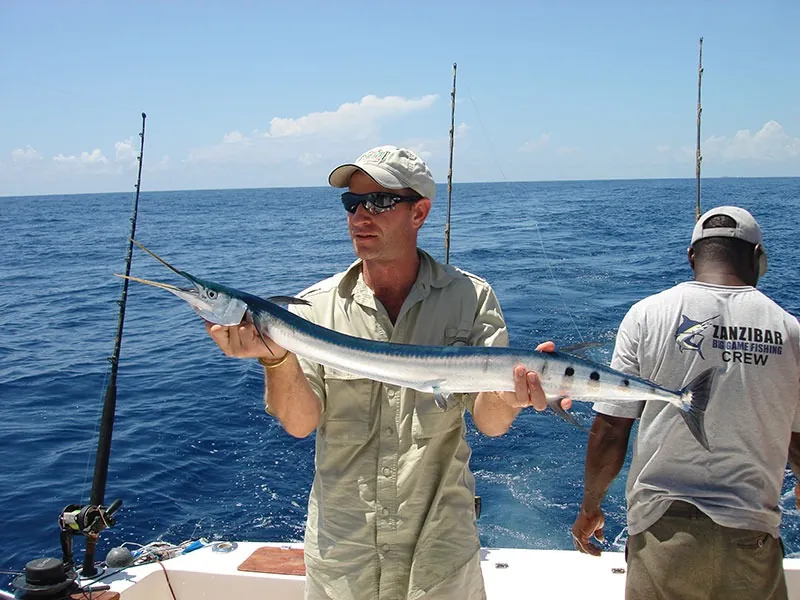 An angler on a boat holding a long needlefish-like fish, crew member behind