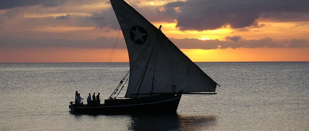 A dhow with a dark lateen sail and passengers silhouetted against an orange sunset