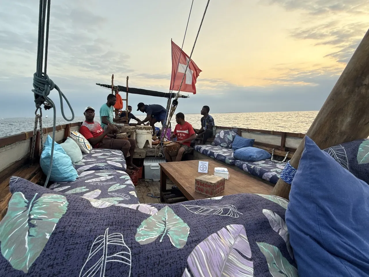 Cushioned seating on a dhow deck at dusk with crew preparing food under a red flag