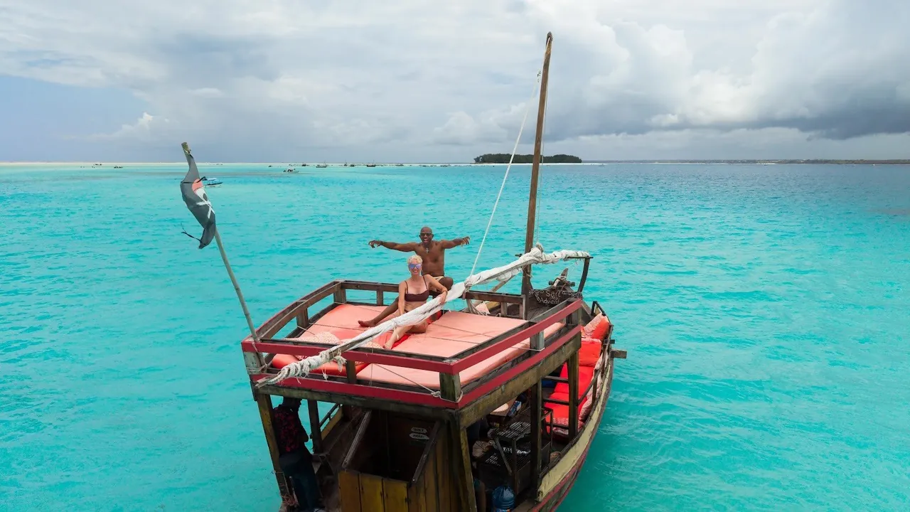 A dhow with a red-cushioned sun deck anchored in bright turquoise water with a low island on the horizon
