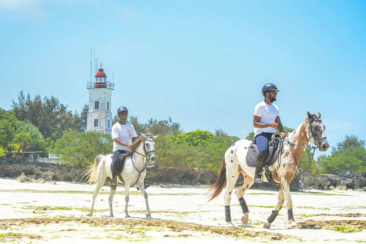 Two riders in helmets on a white sand beach below a red-and-white lighthouse