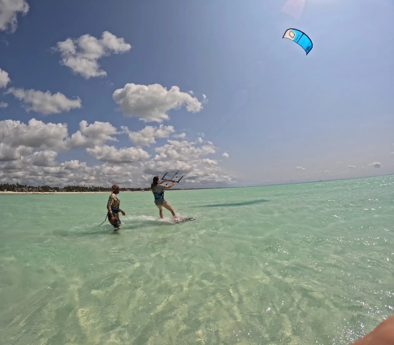 An instructor walking alongside a student riding a kiteboard in knee-deep clear water