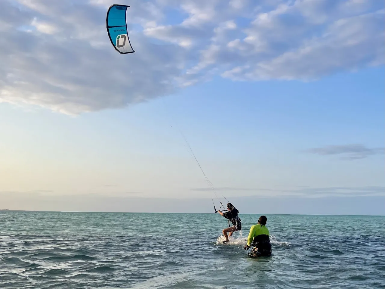 A rider and an instructor in the water at golden hour, with a kite high overhead