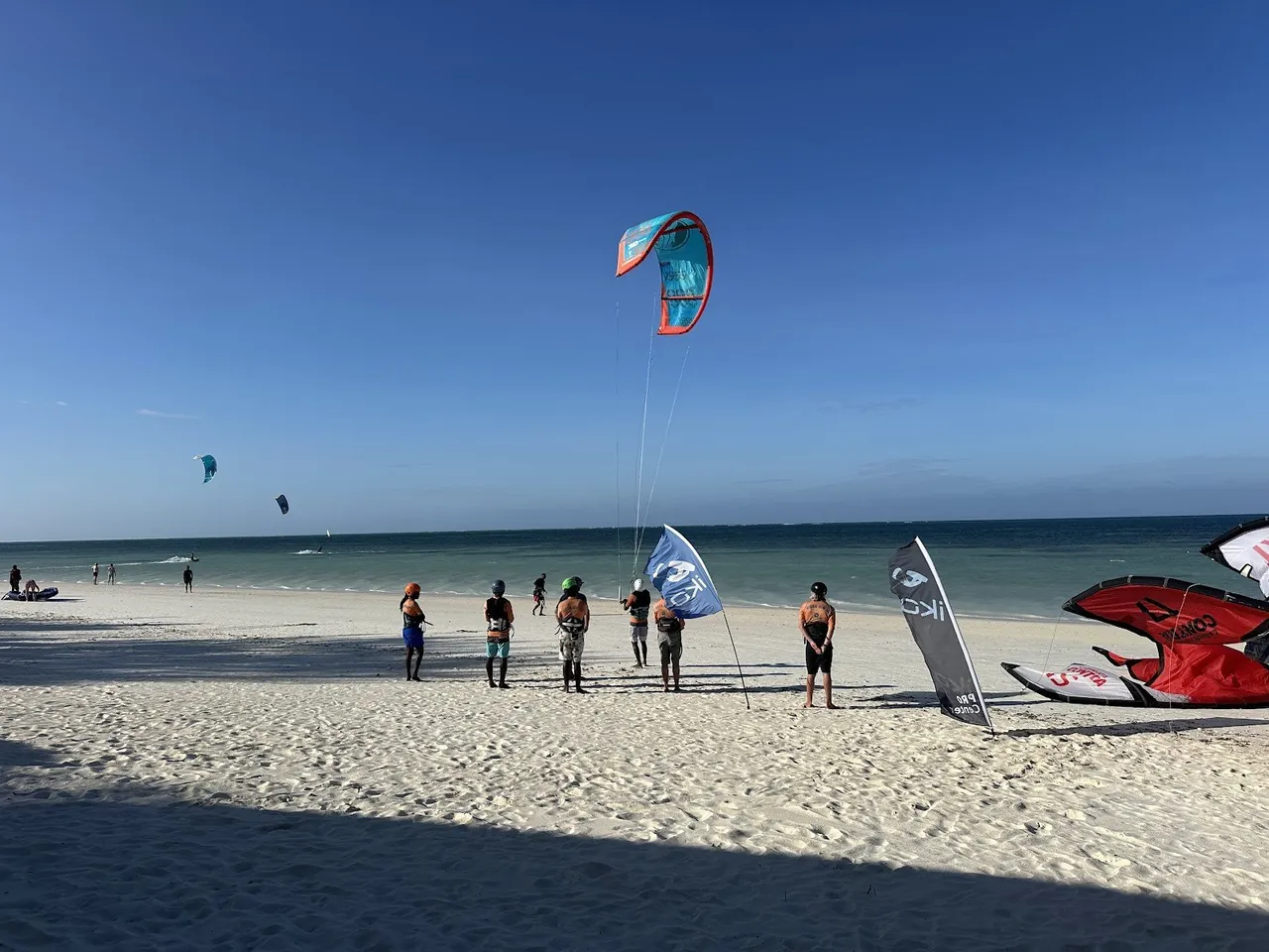 Students in helmets on a white beach with IKO flags and a rigged kite