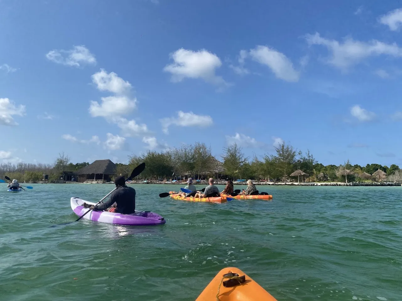 Kayakers paddling on open green water toward a thatched beachfront