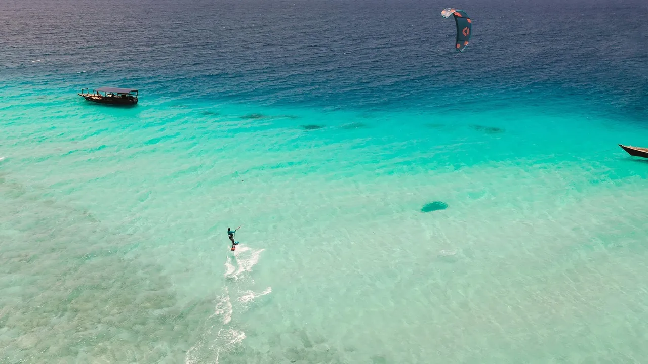 Overhead view of a kitesurfer on bright turquoise shallows with a moored wooden boat