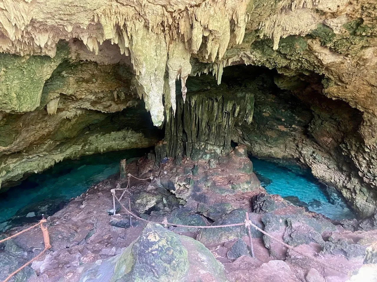 Limestone cave with hanging stalactites above two turquoise pools and a roped path