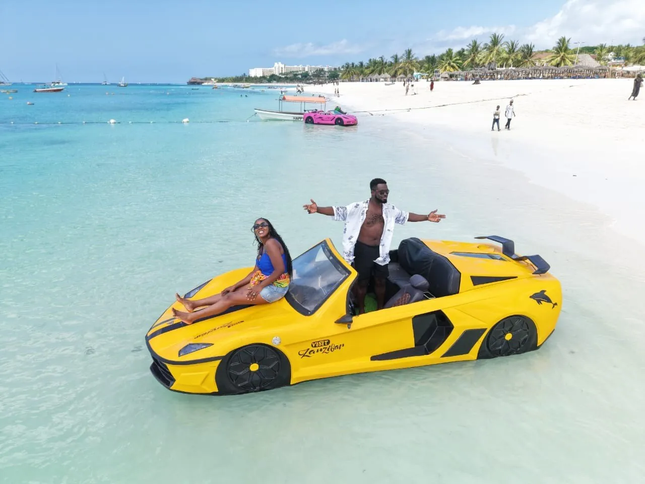 Two people posing on a yellow sports-car-shaped jet car in shallow water off a busy beach