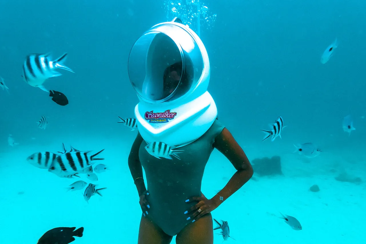 A woman in a swimsuit and air-fed helmet standing on sand with fish around