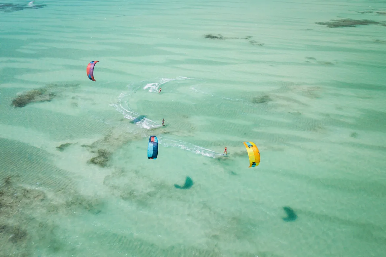 Overhead view of three kitesurfers carving across pale green shallow water