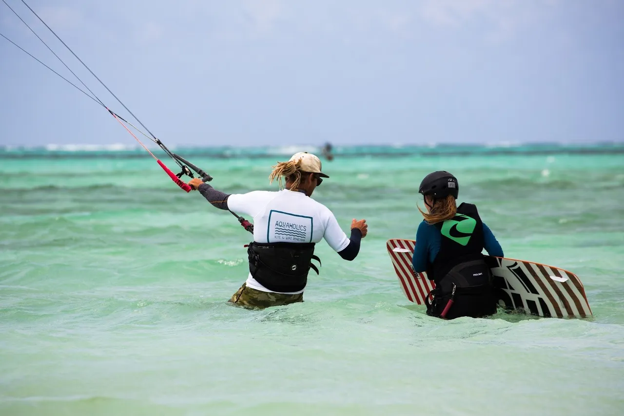 An instructor and a student with a board standing waist-deep in turquoise water, kite lines overhead