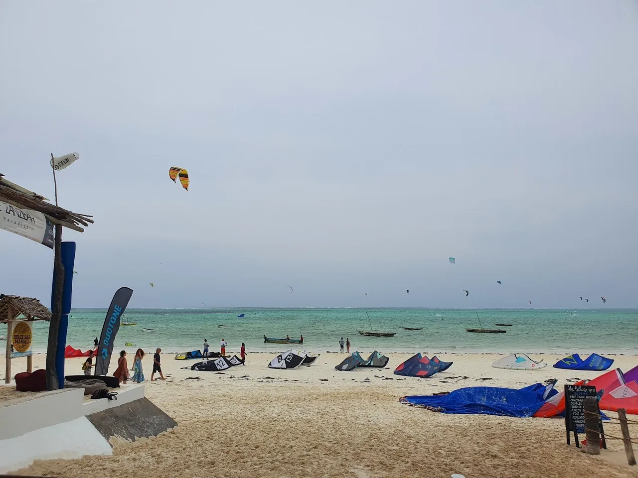 Kites laid out on the sand beside a beach bar with flags, with many kites flying far out over the lagoon