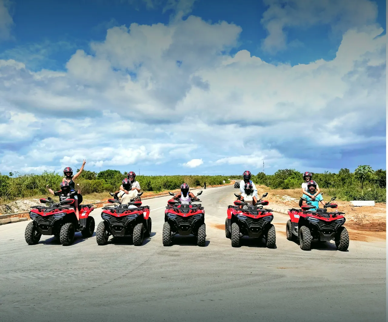Five red quad bikes with riders lined up across a tarmac road under a big sky