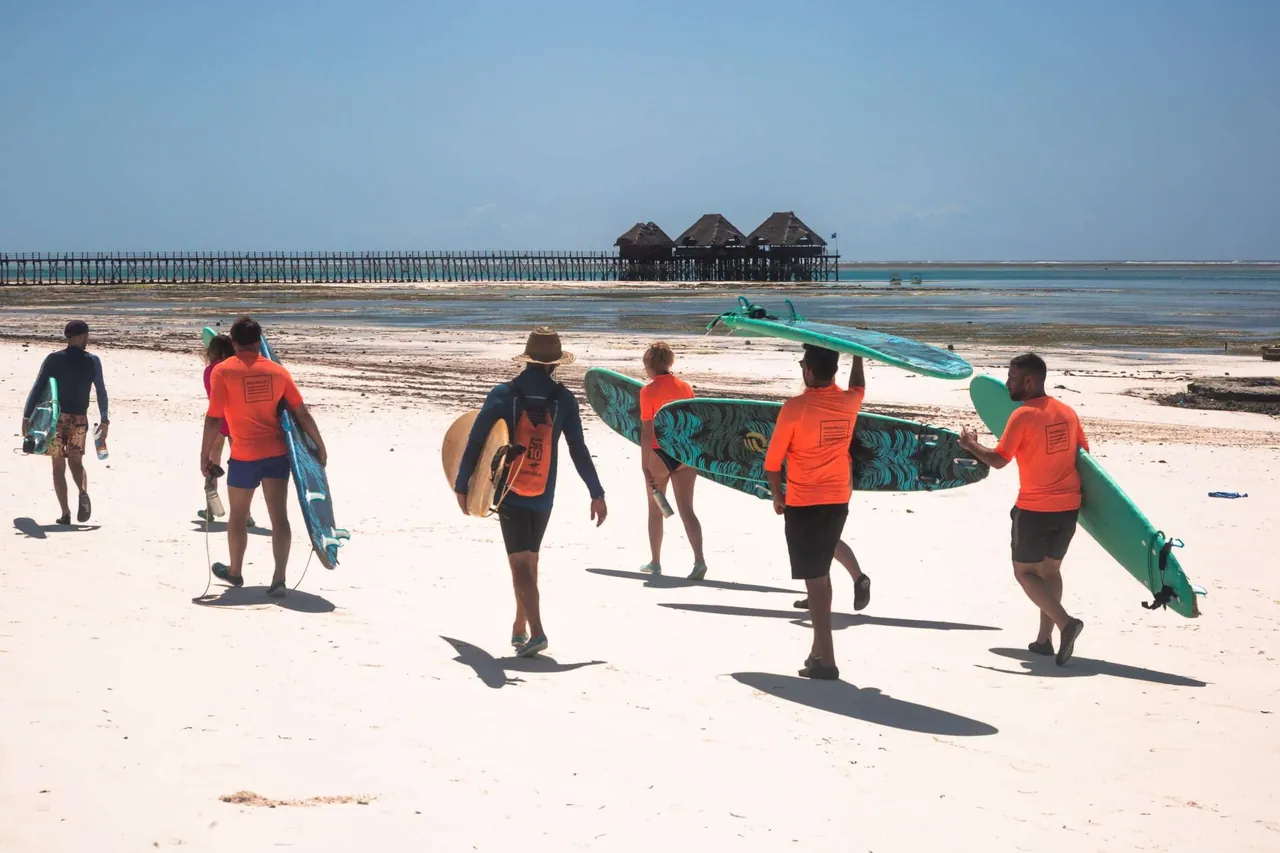 Surfers carrying boards along a white beach toward a long pier with thatched huts