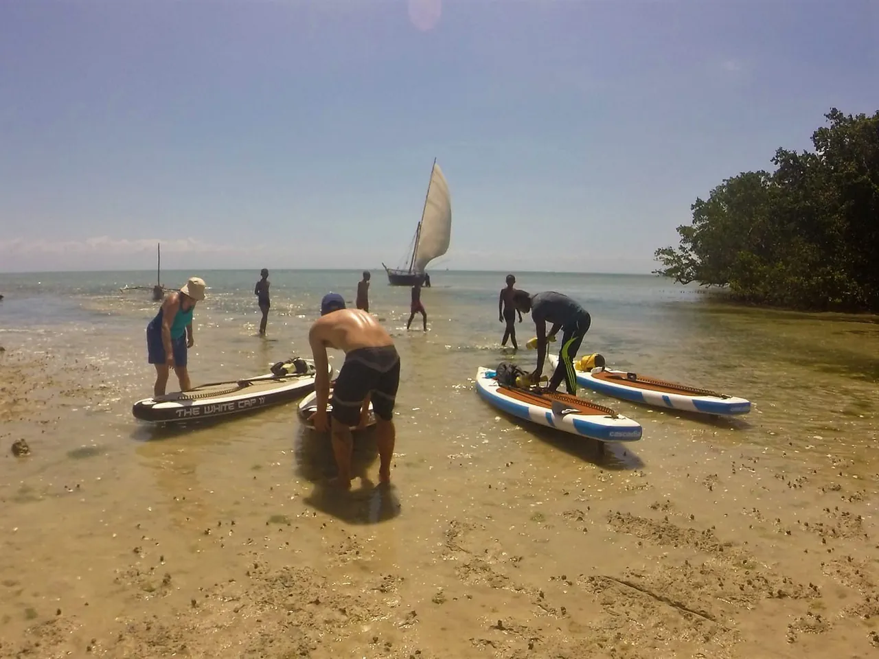People preparing paddleboards on a sandy shore at low tide, with a dhow sail offshore