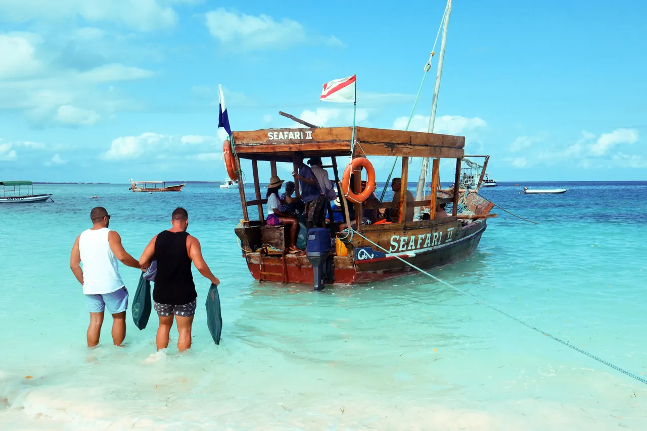 Two men wading out to a wooden dhow named Seafari in turquoise shallows