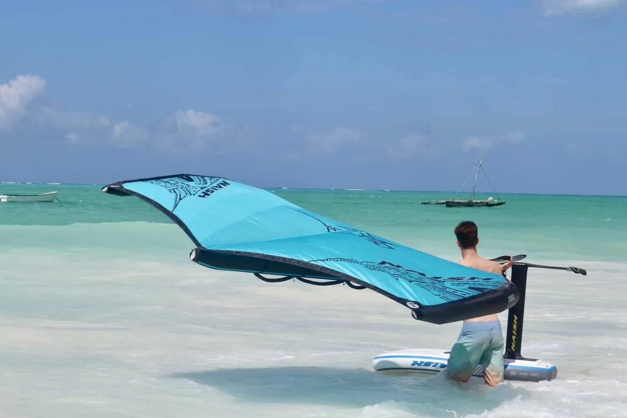 A man kneeling in shallow water holding a blue wing above a board, with a dhow behind