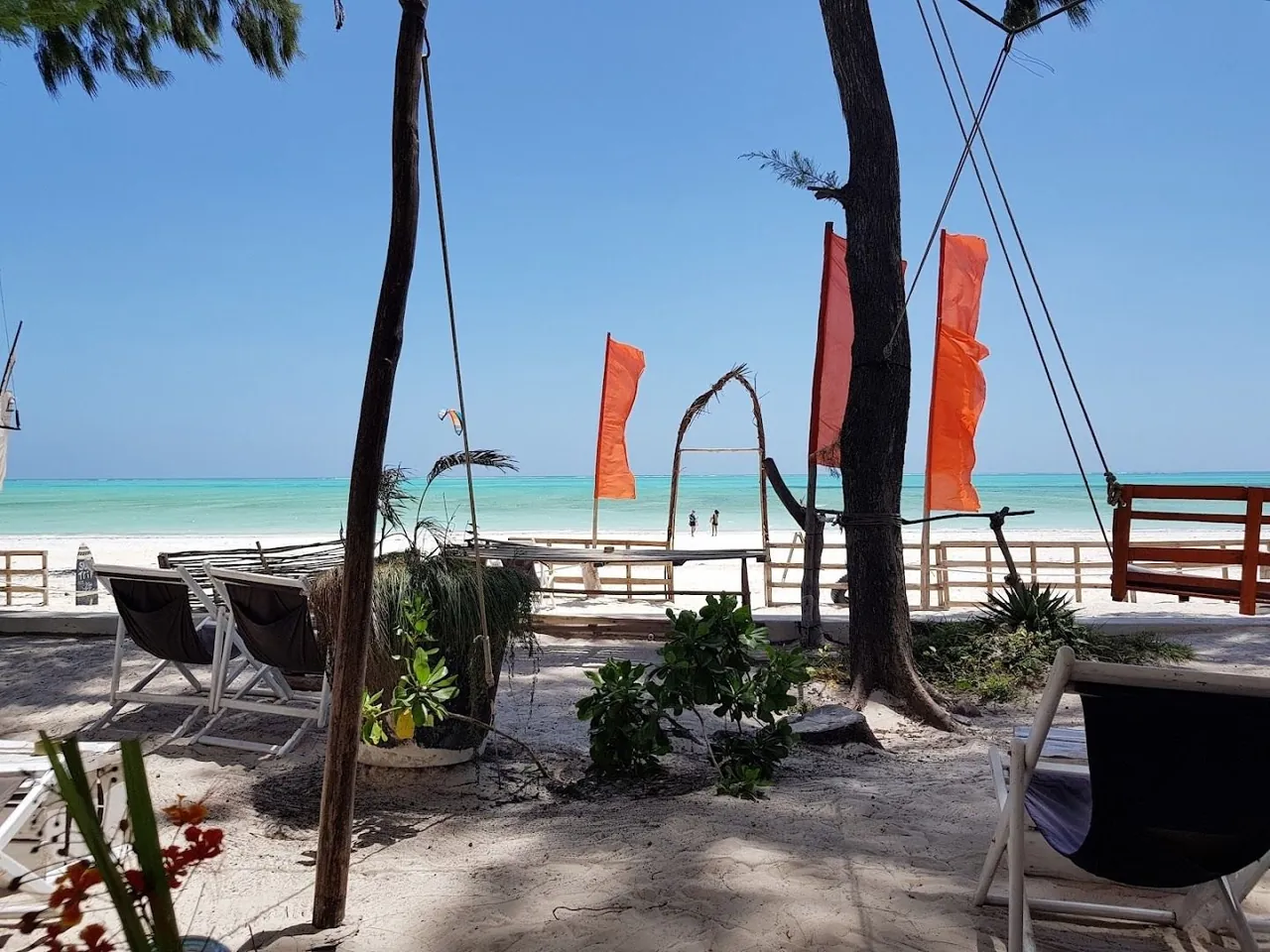 Daybeds and swing seats under trees on white sand, orange flags beside them and a turquoise lagoon beyond.