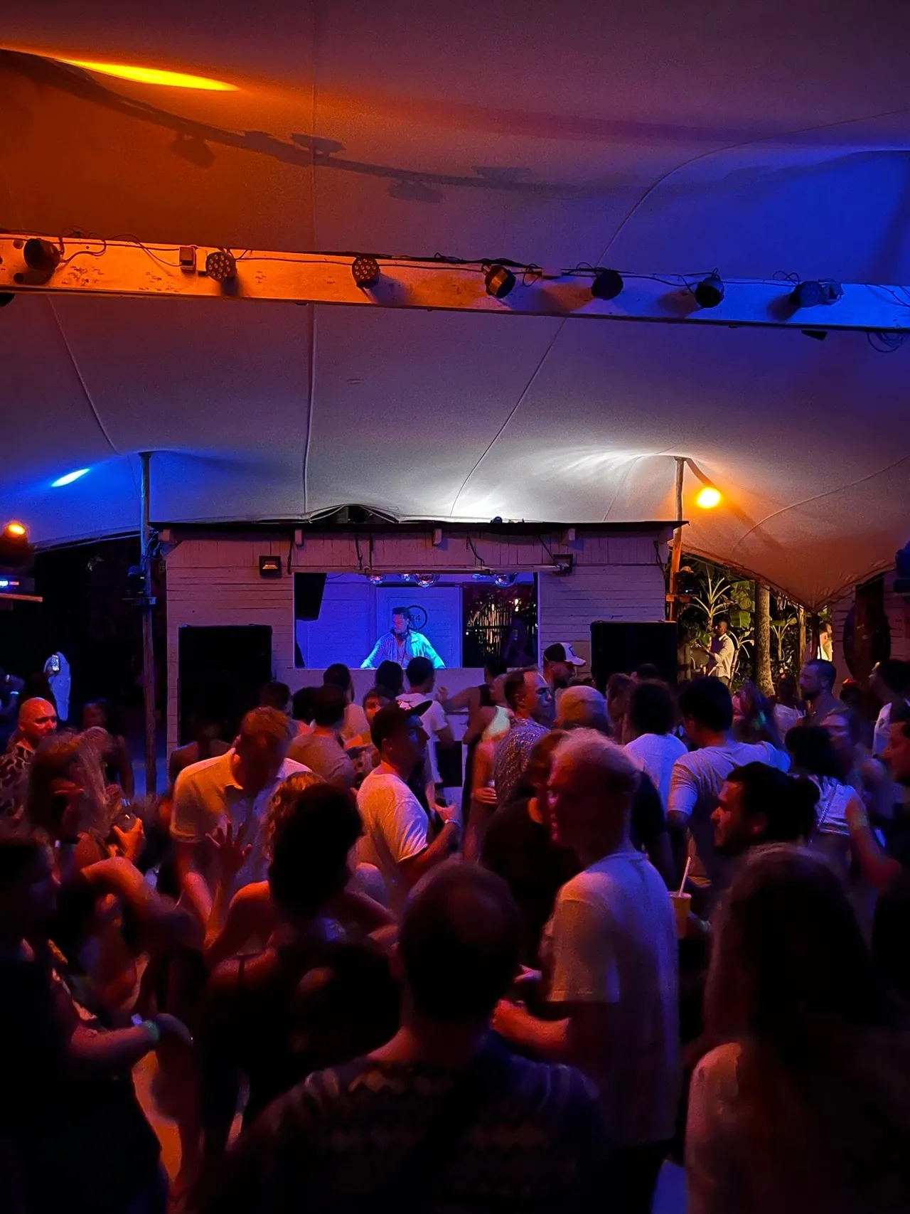 A crowd seen from behind, dancing under a lit canopy in front of a DJ booth at night.