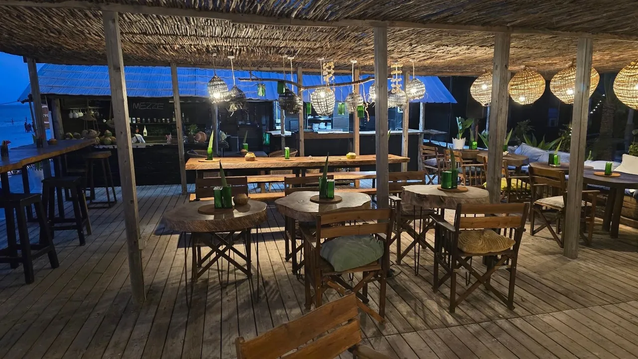 A covered wooden deck with tables, chairs and hanging lamps under a palm-thatch roof.