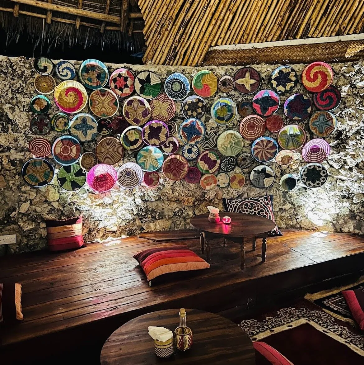 A low lounge platform with cushions and small tables in front of a stone wall covered in colourful woven plates.