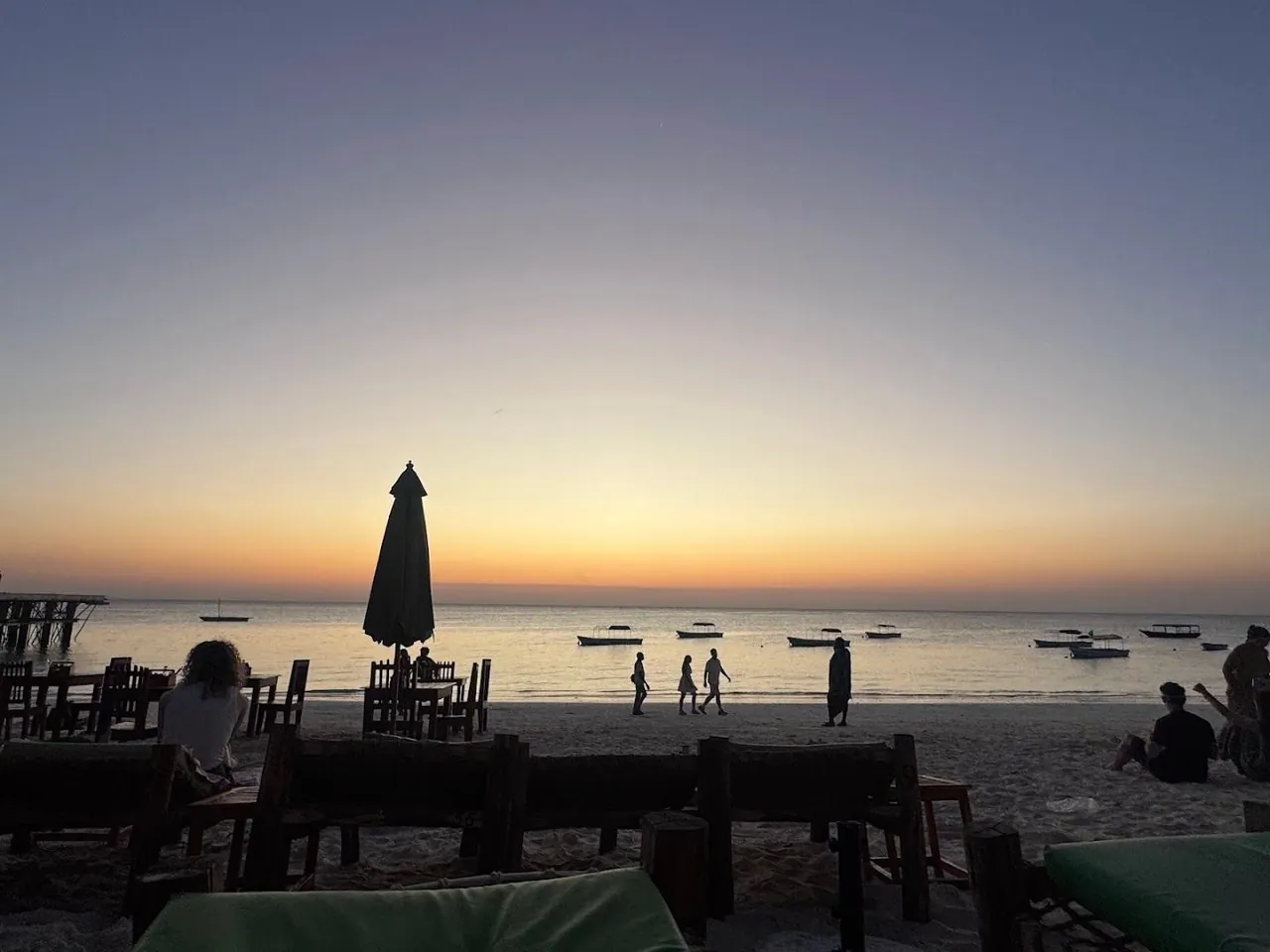 Silhouetted beach tables and a closed parasol at dusk with boats on the sea