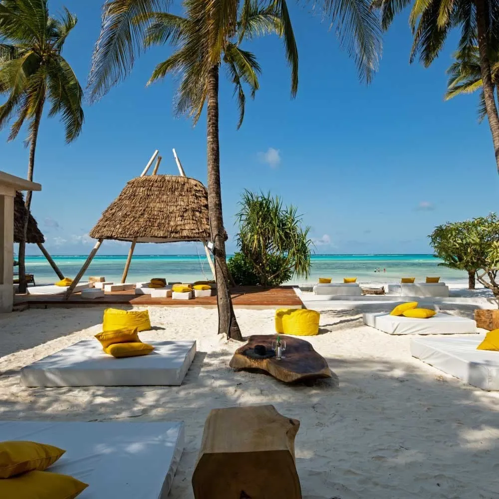 White daybeds with yellow cushions and beanbags under palm trees, a thatched parasol and a turquoise lagoon behind.