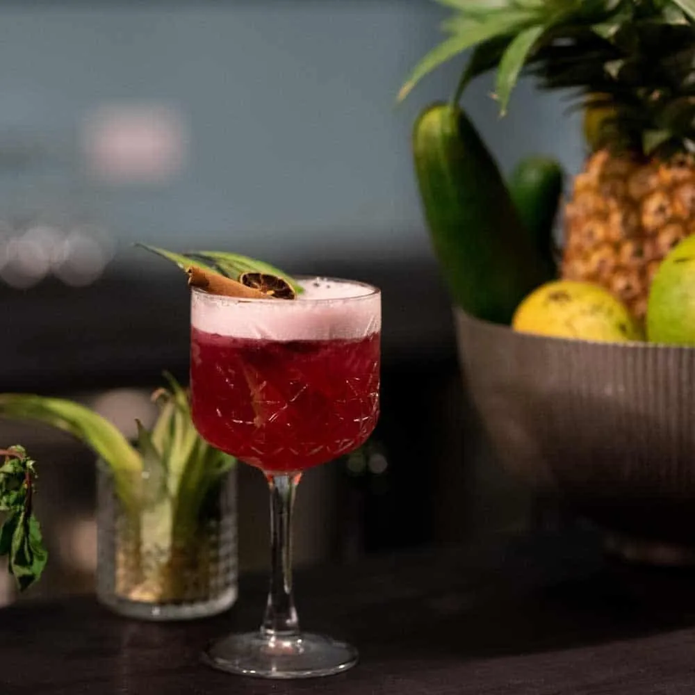 A deep red cocktail garnished with a leaf on a dark table, with a pineapple and fruit bowl behind.