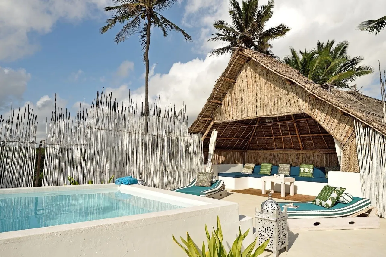 A small plunge pool beside a thatched cabana with blue and white cushions and a reed fence.