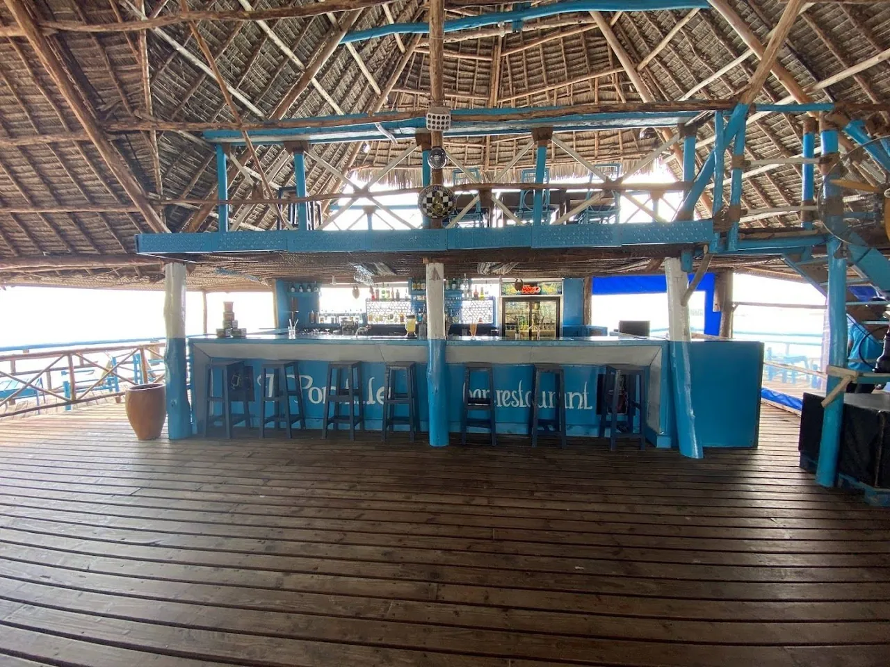 A bar counter painted blue under a high thatched roof on a wooden deck, with the sea visible on both sides.