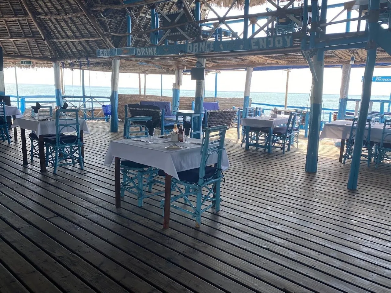 Tables with white cloths and blue chairs on a wooden deck under a thatched roof, open to the sea.