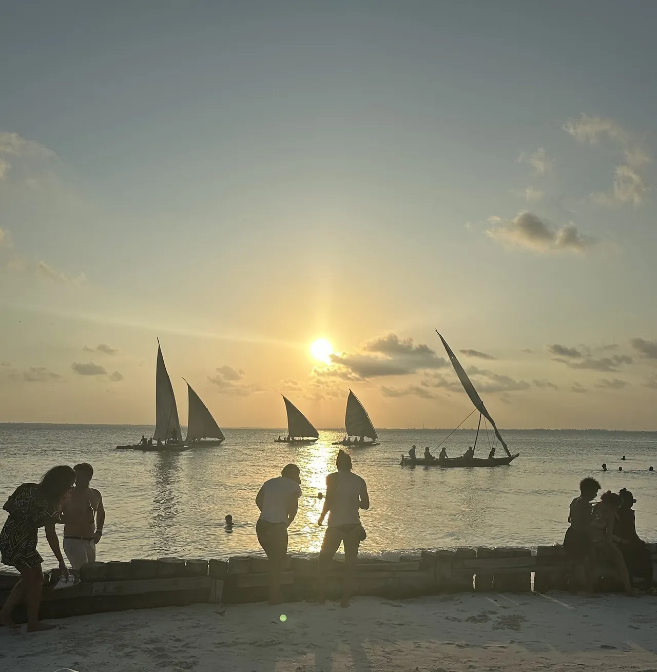 Several dhows sailing past at sunset while people sit along a low wall on the beach.