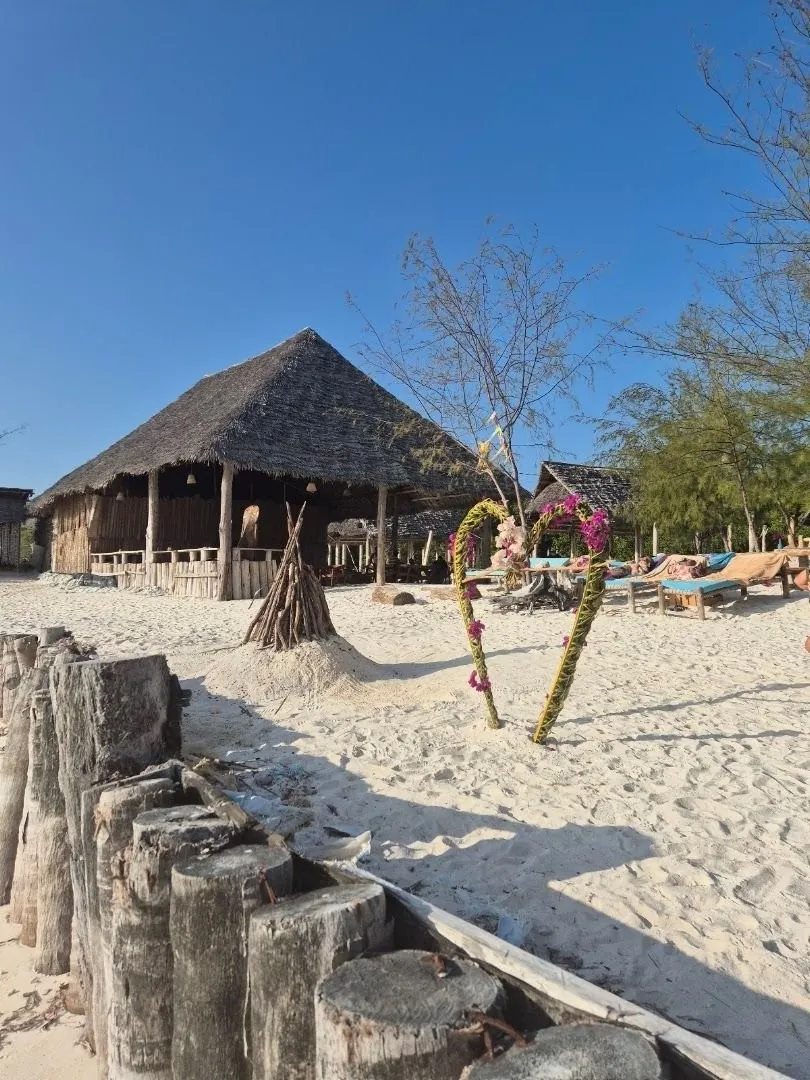 A thatched beach bar hut on white sand with painted driftwood sculptures and a row of wooden posts in front.