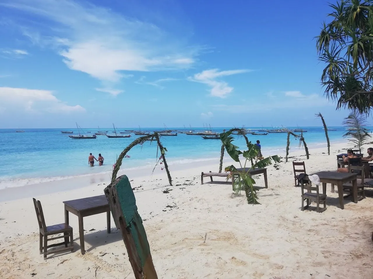Tables and chairs on white sand facing a turquoise sea with fishing boats