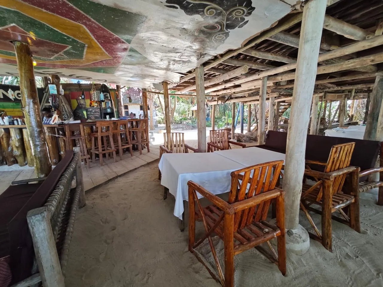 A sandy-floored bar with painted ceiling, wooden tables and a long counter