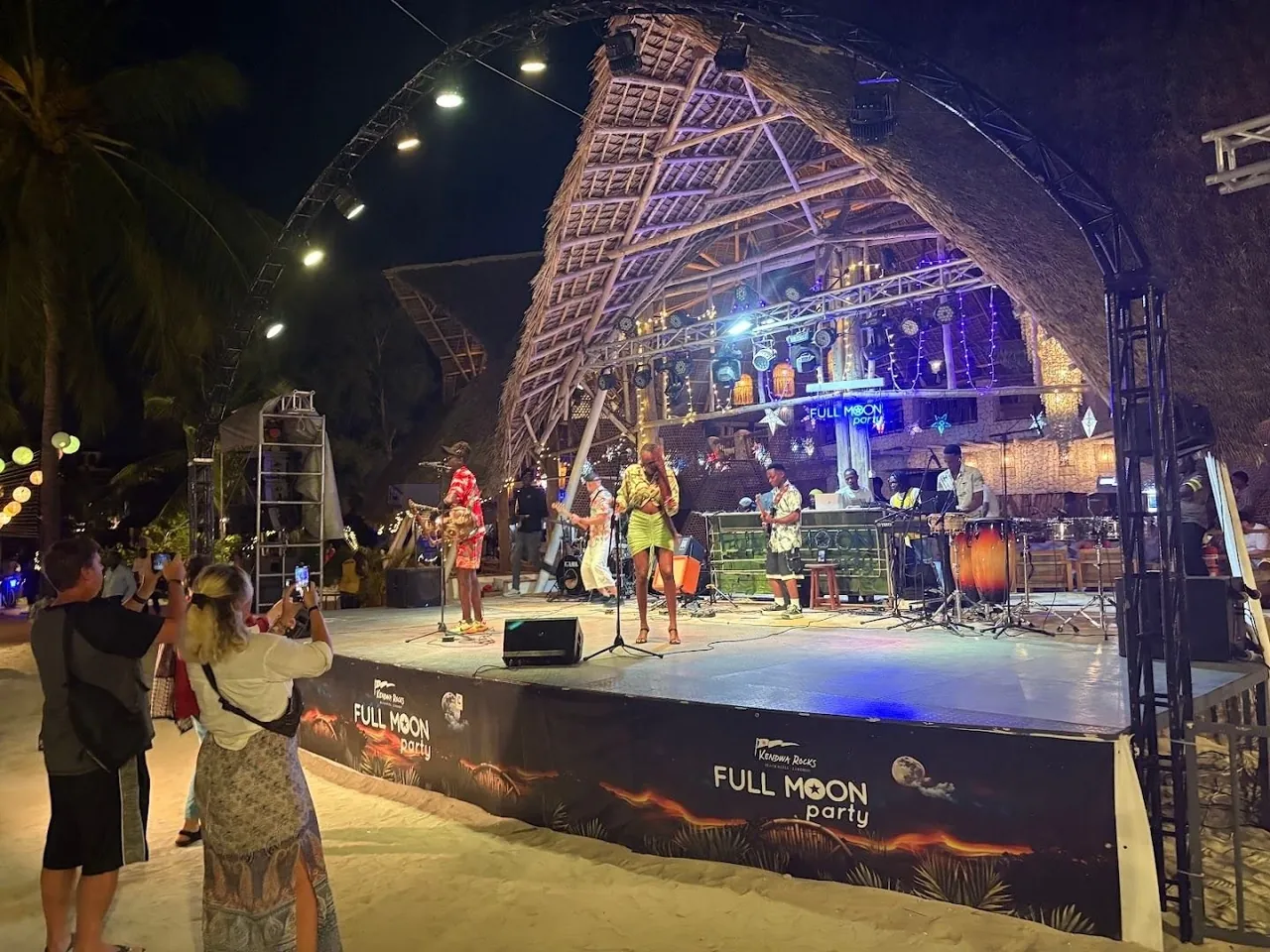 A lit stage under a thatched arch with a band playing and a Full Moon Party banner
