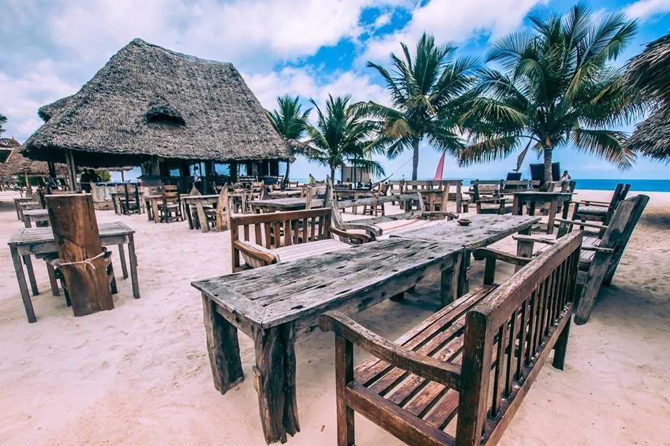 Rustic wooden tables and benches on sand in front of a thatched bar and palms