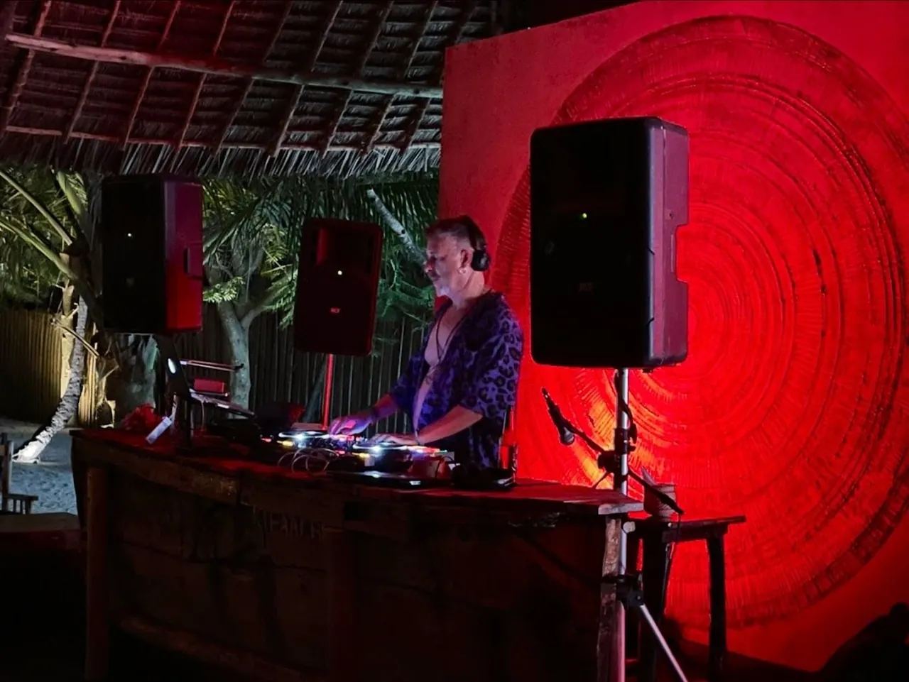 A DJ at decks between speakers in front of a red lit wall
