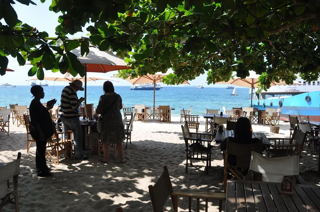 Tables and chairs on the sand under a shady tree, with parasols and the sea beyond
