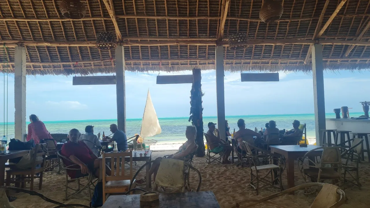 An open-sided restaurant under a thatched roof with diners at tables and a dhow sail visible on the turquoise sea.
