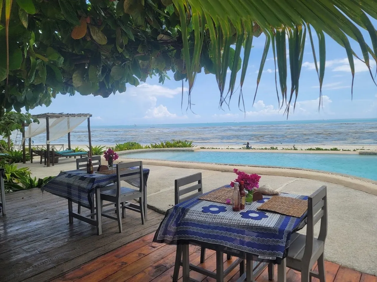 Tables with blue-and-white cloths on a deck beside a pool, framed by palm leaves, with the beach beyond.