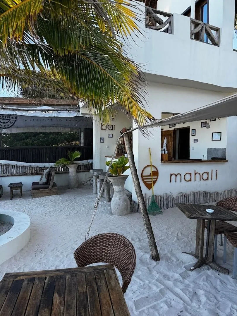 A small white bar kiosk with a "mahali" sign, rattan chairs and tables on raked sand.
