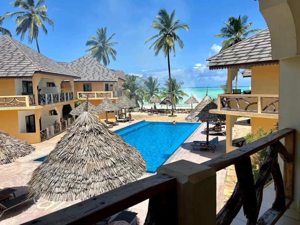 A swimming pool between two-storey buildings, with a thatched parasol in front and palms and the sea beyond.