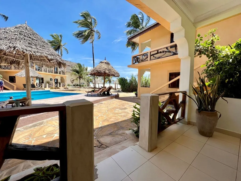 A view from a covered terrace over a pool, thatched parasols and palms towards the beach.