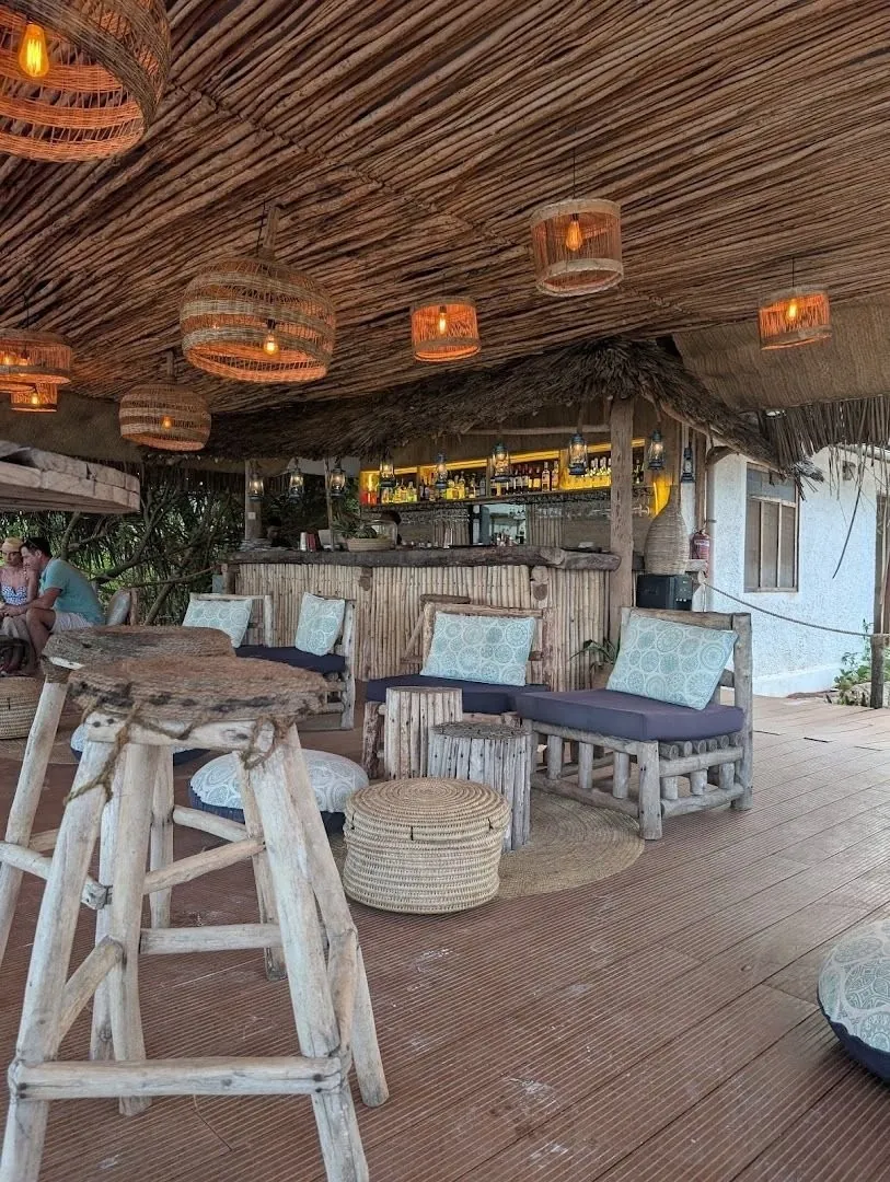 A bar counter under a woven thatched ceiling hung with wicker lamps, with wooden bar stools on a deck.