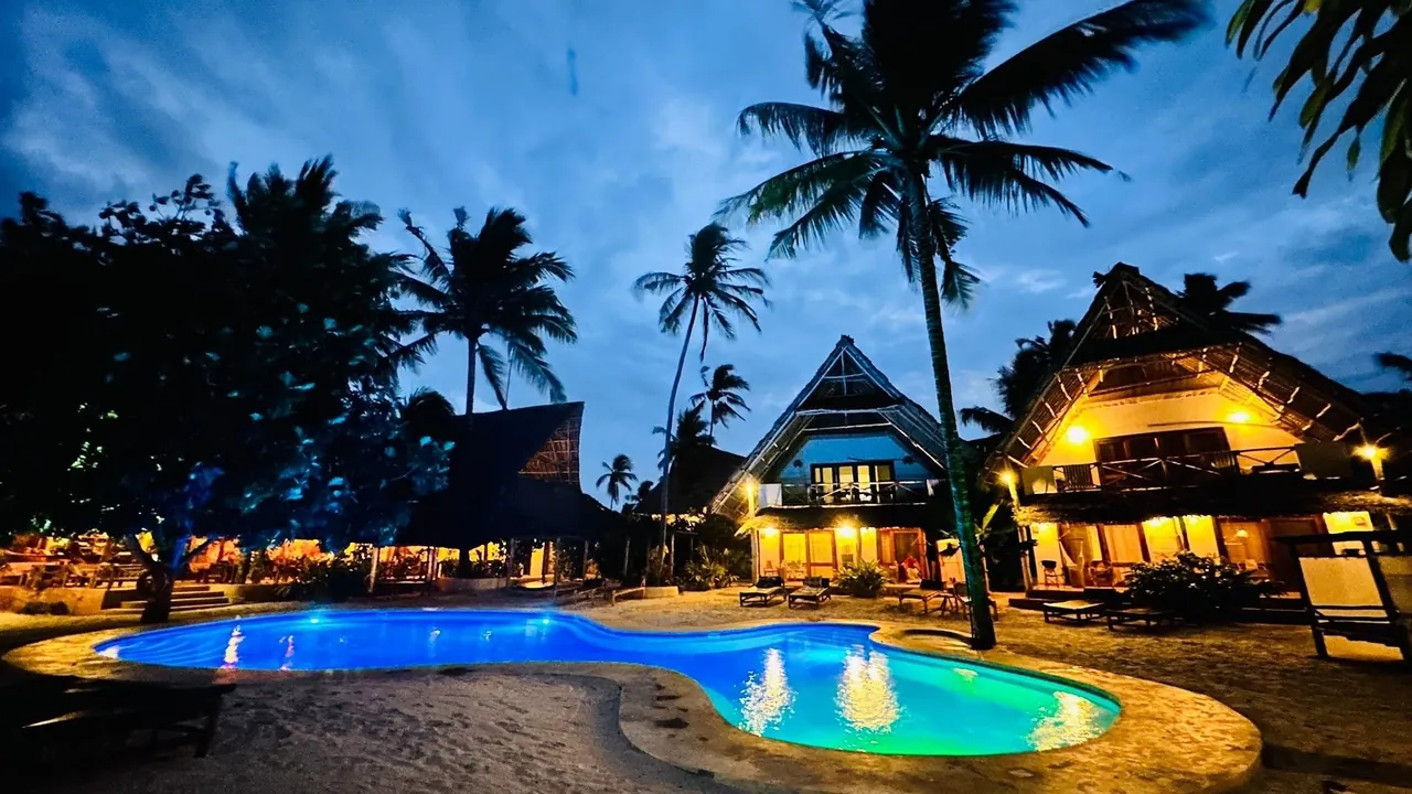 A lit swimming pool at dusk with thatched buildings glowing behind it and palm trees against a blue evening sky.