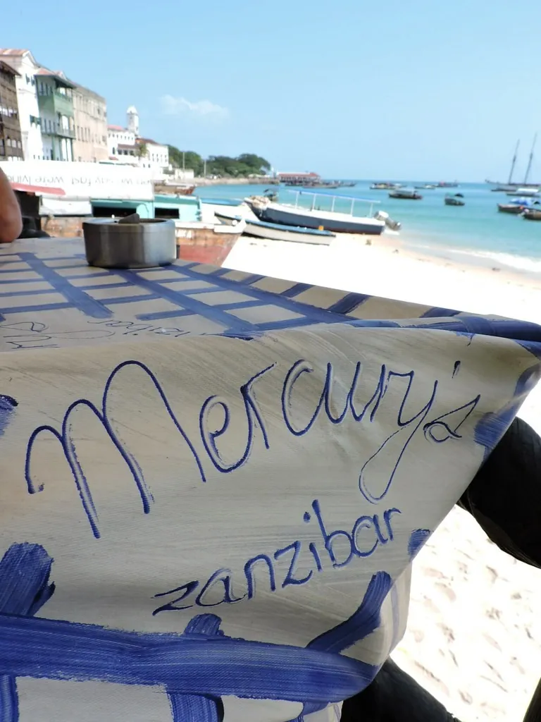 A table with a hand-painted Mercury's cloth on the sand, with Stone Town's beach and boats behind