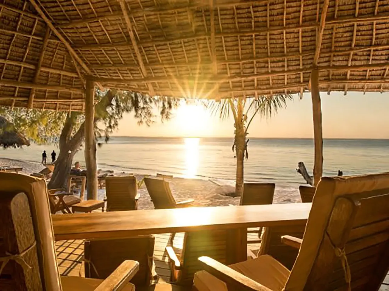 The sun setting over calm water, seen from wooden chairs and a table under a thatched roof.